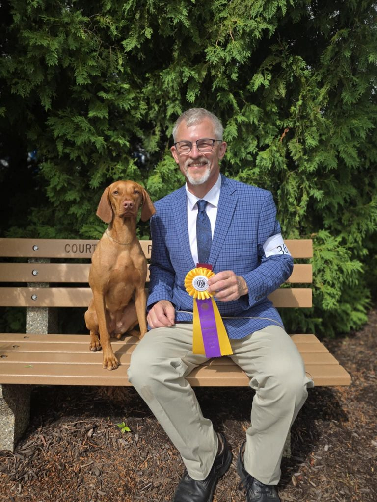 Mabel, a red Vizsla, sitting up on a park bench beside her smiling handler, who holds the purple and gold rosette she won.