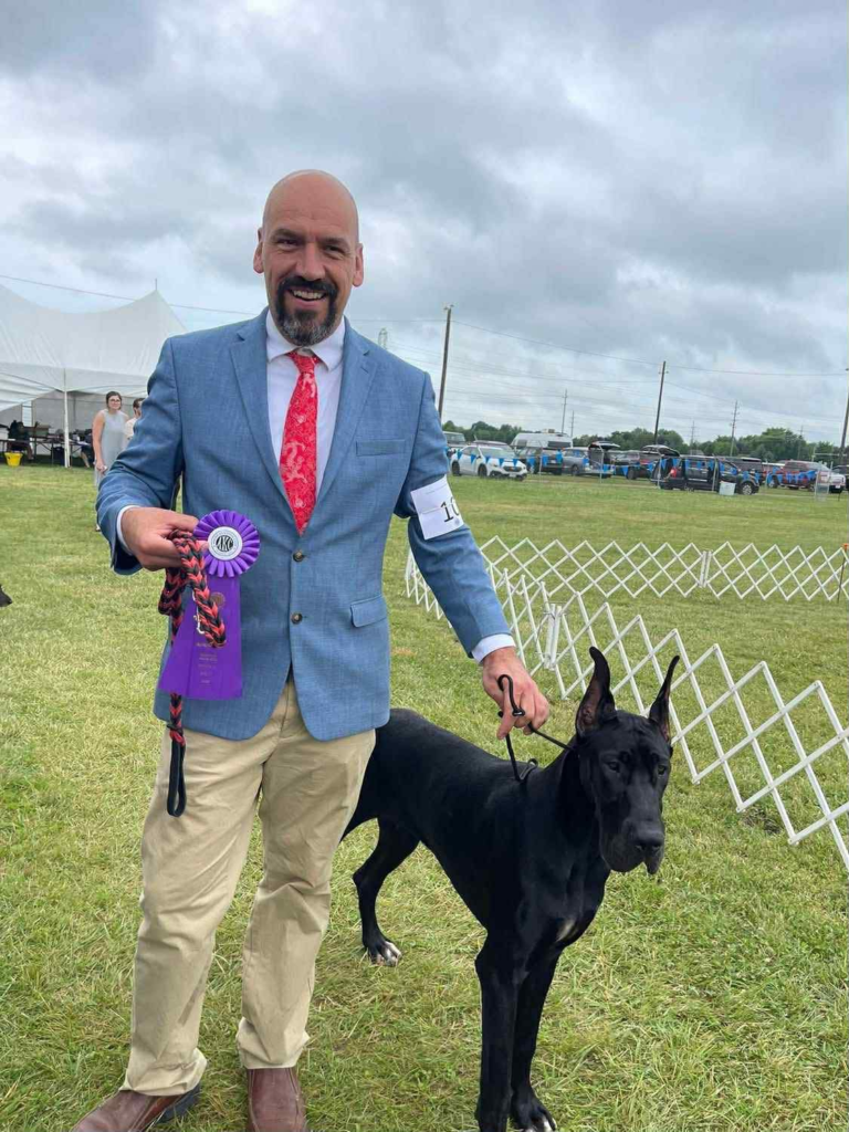 Mark smiling in a blue show jacket, holding a purple AKC rosette, with Clementine, a young black Great Dane, at his side on the show grounds.