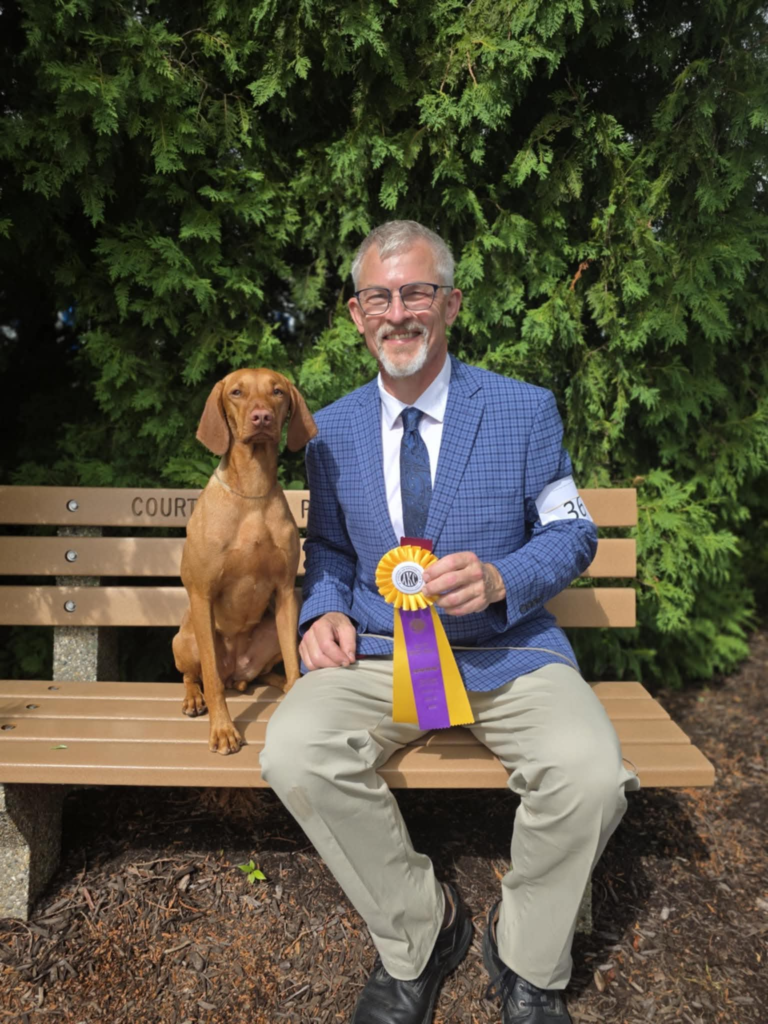 Mike smiling on a park bench in a blue checked jacket, holding a purple and gold AKC rosette, with Mabel, a red Vizsla, sitting up beside him.
